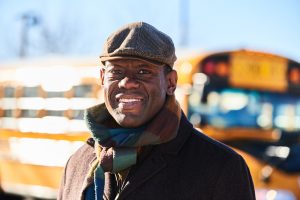 A Black man stands outside, dressed in winter clothes, with a yellow school bus behind him.