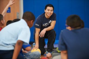An Asian American man kneels on the floor of a school gym with several elementary students sitting around him.
